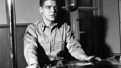 Radio-DJ Nick Clooney sitzt an einem Mikrofon und einem Plattenspieler im AFN-Studio in Frankfurt, Schwarz-weiß-Aufnahme aus den 1950er Jahren