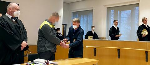Foto einer Gerichtsverhandlung. Ein Justizbeamter nimmt dem Angeklagten die Handschellen ab. Die Richter nehmen Platz.