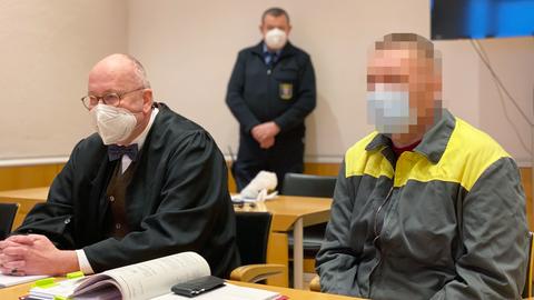 Ein Angeklagter im Gerichtssaal, neben seinem Anwalt sitzend. Im Bildhintergrund unscharf ein Justizvollzugsbeamter.