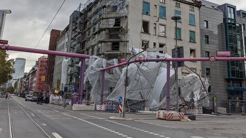 An einer Straßenmündung ist die Hälfte eines Gerüsts nach unten abgerutscht. Eine weiße Plane weht aus den Gerüst-Stäben heraus. Im Vordergrund sind rosafarbene Rohre zu sehen, die hier oberirdisch über die Straße geleitet werden.