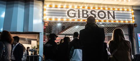 Mehrere Menschen stehen in einer Warteschlange vor einem Eingangstor, darüber steht in leuchtender Schrift der Name "Gibson".