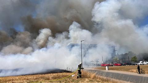 Feuerwehrmann mit Schlauch auf Feld, viel Rauch, im Hintergrund Gebäude mit Autos davor
