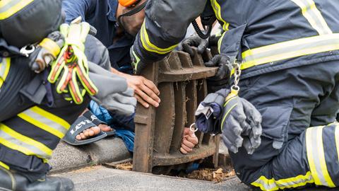 Feuerwehrleute um Mann, dessen Hand im Kanaldeckel feststeckt.