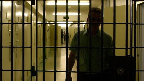Gitterstäbe in einer Haftanstalt, dahinter ein Vollzugsbeamter