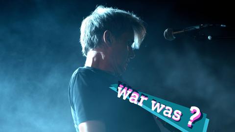 Foto: Dirk von Lowtzow am Mikrofon während eines Konzertes, umgeben von Licht und Nebel. Auf dem Bild eine kleine, farbige Grafik mit dem Schriftzug "war was?".