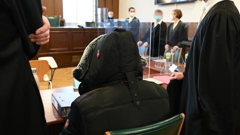Foto des Gerichtssaals: im Bildvordergrund sitzt ein Mann in Jacke mit hochgezogener Kapuze, daneben zwei Anwälte. Im Bildhintergrund stehen weitere Anwälte und Richter (unscharf).