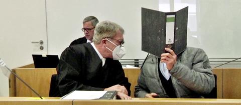 Ein Mann hält sich in einem Gerichtssaal einen Aktenordner vors Gesicht, während er mit einem Anwalt spricht.