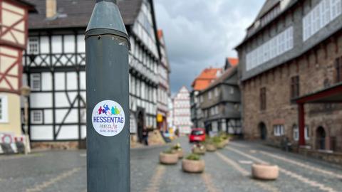 Ein Aufkleber vom Hessentag klebt an einem Laternenmast. Im Hintergrund ist rechts das Rathaus und mehrere Fachwerkhäuser zu sehen.