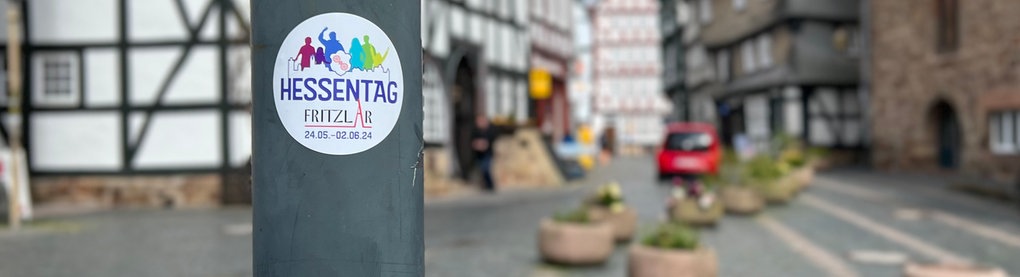 Ein Aufkleber vom Hessentag klebt an einem Laternenmast. Im Hintergrund ist rechts das Rathaus und mehrere Fachwerkhäuser zu sehen.