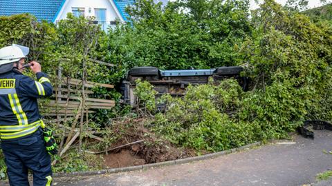 Ein Auto liegt auf dem Dach in einer grünen Hecke. Ein Feuerwehrmann steht daneben.