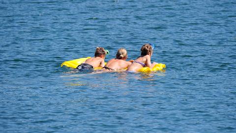 Three young men lie face down on an air mattress floating in a lake.