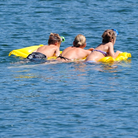 Three young men lie face down on an air mattress floating in a lake.