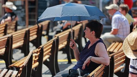 Eine Frau sitzt auf einer Bank und hält einen Schirm als Sonnenschutz über sich.