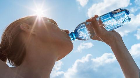 Eine Frau trinkt aus einer Wasserflasche. Sie wurde von unten gegen den Himmel fotografiert, so dass sie gleißende Sonne Frau und Flasche etwas überstrahlt.