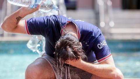 Ein Mann in blauem T-Shirt sitzt auf dem Rand eines Brunnens und schüttet sich Wasser aus einer Flasche über den Kopf.