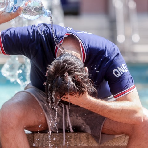 Ein Mann in blauem T-Shirt sitzt auf dem Rand eines Brunnens und schüttet sich Wasser aus einer Flasche über den Kopf.