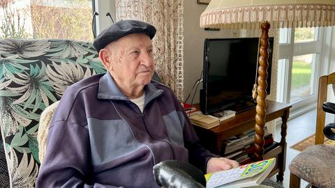 Ein sehr alter Mann sitzt in einem Sessel und hält ein Rätselheft in der Hand.