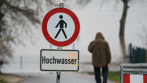 Ein Mann geht hinter einem Warnschild auf dem "Hochwasser" steht zum Rheinufer.