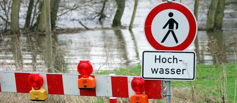 Ein über die Ufer getretener Fluss. Davor ein Schild "Hochwasser" und ein rot-weißes Absperrgestell.