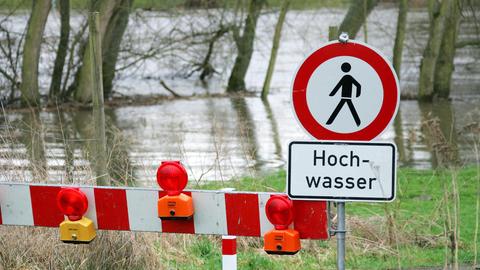 Ein über die Ufer getretener Fluss. Davor ein Schild "Hochwasser" und ein rot-weißes Absperrgestell.