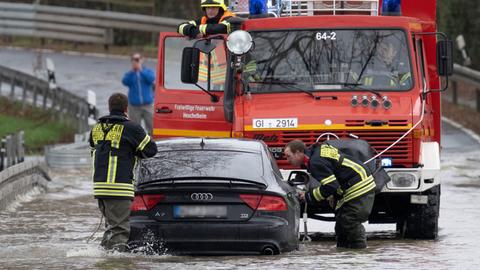 Ein Auto steht mit den Reifen im tiefen Wasser. Feuerwehrleute sind mit einem Einsatzauto vor Ort.