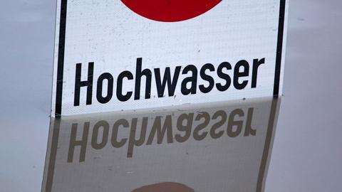 Nahaufnahme eines Schildes, das im Wasser steht: Das Wort "Hochwasser" spiegelt sich im grauen Wasser, das das Schild umgibt.