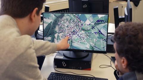 Ein Schüler zeigt auf den Bildschirm eines Rechners, auf welchem eine fotorealistische Simulation von Hochwasser in einer Ortschaft zu sehen ist.