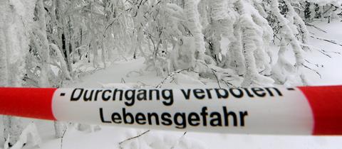 "Durchgang verboten - Lebensgefahr" steht auf einem rot-weißen Absperrband vor einer schneebedeckten Straße.