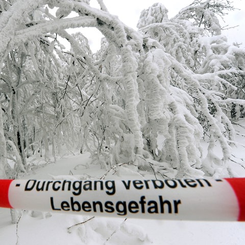 "Durchgang verboten - Lebensgefahr" steht auf einem rot-weißen Absperrband vor einer schneebedeckten Straße.