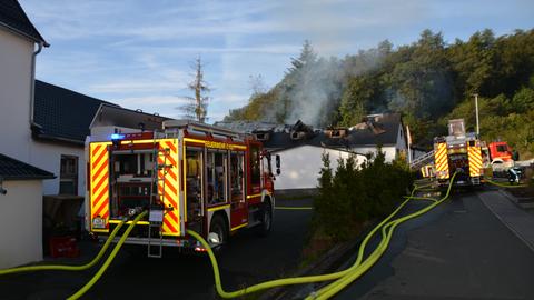 Feuerwehrwagen mit Feuerwehrschläuchen an weißem Gebäude, dessen Dachstuhl sichtbar beschädigt ist und aus dem es raucht.