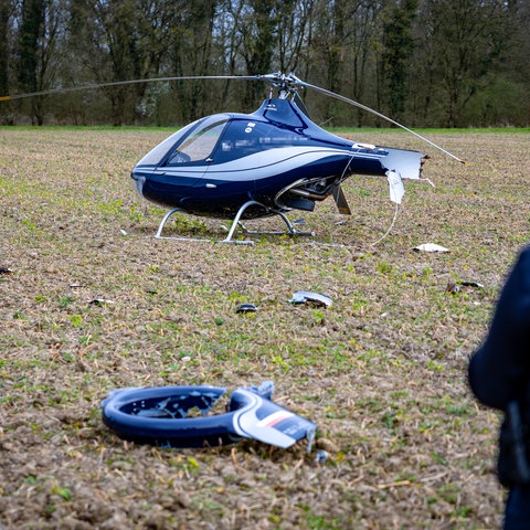 Das Bild zeigt einen abgestürzten Hubschrauber auf einem Feld. Im Gras liegen abgebrochene Teile. Rechts am Bildrand ist unscharfe Polizistin. 