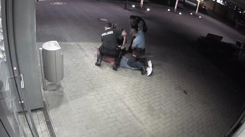Drei Menschen in Polizeiuniformen knien auf einer Person, die auf dem Boden liegt. Ein vierter steht daneben.