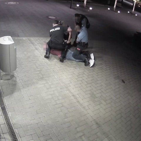 Drei Menschen in Polizeiuniformen knien auf einer Person, die auf dem Boden liegt. Ein vierter steht daneben.