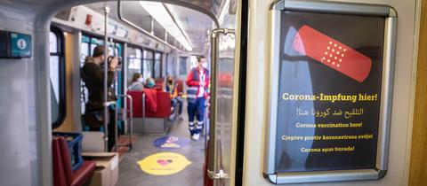 Blick ins Innere einer U-Bahn, in der ein Mensch mit medizinischer Kleidung steht. Im Bildvordergrund ist ein Plakat an der Wand der U-Bahn zu sehen, auf dem in mehreren Sprachen "Corona-Impfung hier" steht.