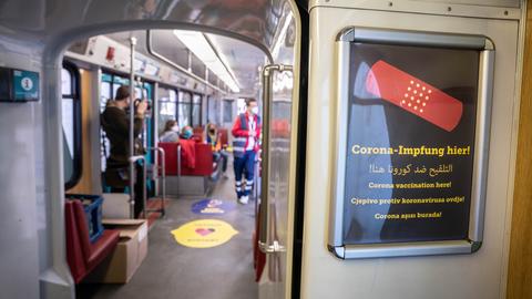 Blick ins Innere einer U-Bahn, in der ein Mensch mit medizinischer Kleidung steht. Im Bildvordergrund ist ein Plakat an der Wand der U-Bahn zu sehen, auf dem in mehreren Sprachen "Corona-Impfung hier" steht.