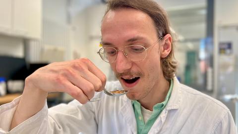 Ein Mann in einem weißen Laborkittel hält einen Löffel und ist kurz davor die Mehlwürmer, die darauf liege zu essen. 