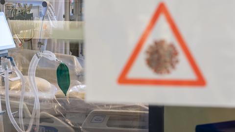 Ein Intensivbett mit Beatmungseinheit steht auf einer Corona-Station hinter einer Scheibe, an der ein Symbol mit einem stilisierten Coronavirus angebracht ist. 