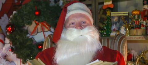 Ein Weihnachtsmann sitzt mit einem dicken Buch in einem Sessel, hinter ihm Tannen und Christbaumkugeln.