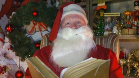 Ein Weihnachtsmann sitzt mit einem dicken Buch in einem Sessel, hinter ihm Tannen und Christbaumkugeln.