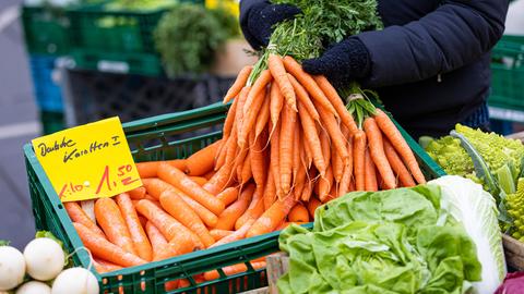 Eine Person mit schwarzen Handschuhen hebt an einem Marktstand ein Bündel Karotten aus einer grünen Kiste hoch.