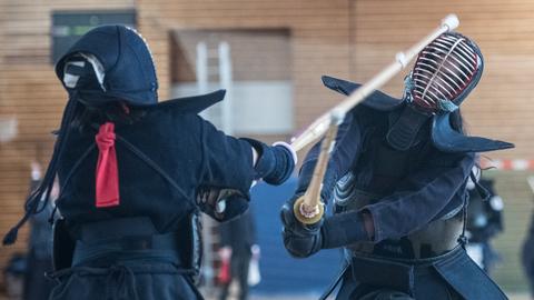 Zwei Menschen in schwarzer Kleidung und mit Gesichtsmasken kämpfen mit Schwertern in einer Sporthalle.