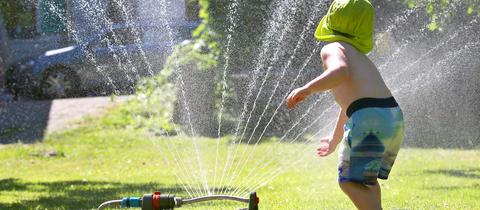 Ein Kind in Badehose und Sonnenhut spielt an einem Rasensprenger auf einem grünen Rasen.