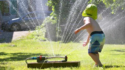 Ein Kind in Badehose und Sonnenhut spielt an einem Rasensprenger auf einem grünen Rasen.