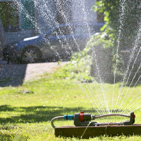 Ein Kind in Badehose und Sonnenhut spielt an einem Rasensprenger auf einem grünen Rasen.