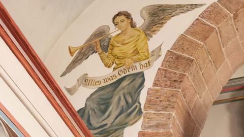 Foto einer Wandmalerei im Innenraum einer Kirche. Auf den Körper eines fliegenden Engels wurde der Kopf der Schauspielerin Jane Russell gemalt.