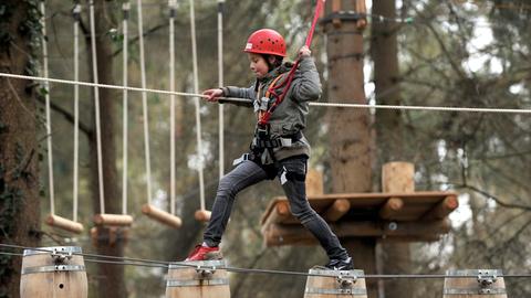 Ein Kind läuft angeseilt über Holzfässer, die zwischen zwei Bäume gespannt sind. 