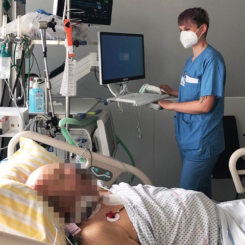Foto: Ein Mann liegt schwerkrank in einem Bett. Daneben viele technische Geräte und eine Pflegerin mit Maske und Arbeitskleidung.