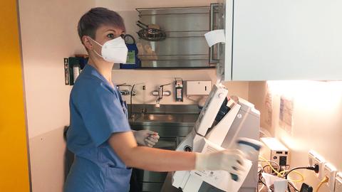 Foto: Ein Pflegerin mit Maske und Arbeitskleidung bedient ein technisches Gerät in einem Arbeitsraum.