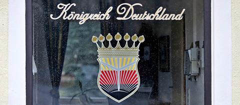 Foto eines Fensters, auf welchem ein Emblem und darüber ein Schriftzug "Königreich Deutschland" in Schreibschrift angebracht ist. Das Emblem zeigt ein Wappen, das eine Krone trägt. Es gliedert sich in schwarz, rot, goldene Flächen (von unten nach oben), wobei sich vom schwarzen "Grund" helle Strahlen nach oben richten.