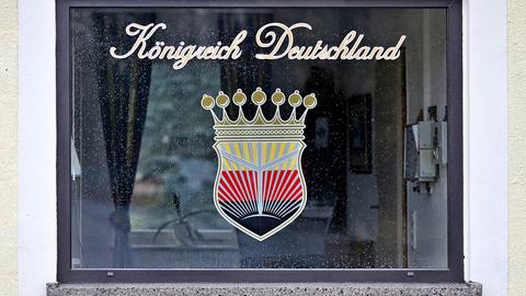 Foto eines Fensters, auf welchem ein Emblem und darüber ein Schriftzug "Königreich Deutschland" in Schreibschrift angebracht ist. Das Emblem zeigt ein Wappen, das eine Krone trägt. Es gliedert sich in schwarz, rot, goldene Flächen (von unten nach oben), wobei sich vom schwarzen "Grund" helle Strahlen nach oben richten.